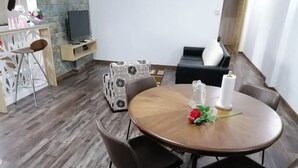 Una sala de estar moderna con una mesa redonda de madera, cuatro sillas negras, un sofá con estampado blanco y negro, un televisor y un jarrón decorativo con una rosa.