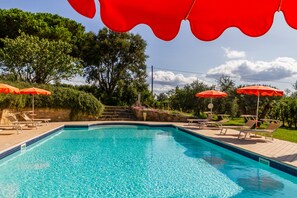 A swimming pool with red umbrellas, lounge chairs, and a stone wall with steps leading up to a grassy area.