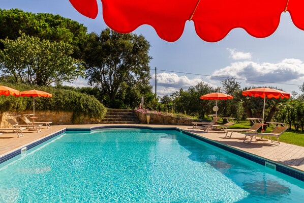 Una piscina con sombrillas rojas, sillones de descanso y un muro de piedra con escalones que conducen a una zona cubierta de césped.