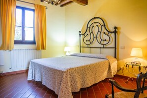 Un dormitorio con cama, una silla, ventana con cortinas, lámpara y radiador.