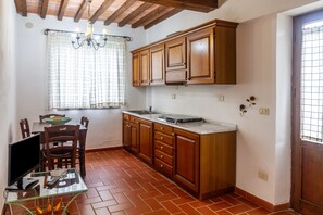 Une cuisine avec des meubles de rangement en bois, une table à manger, des chaises, un téléviseur et une fenêtre avec des rideaux.