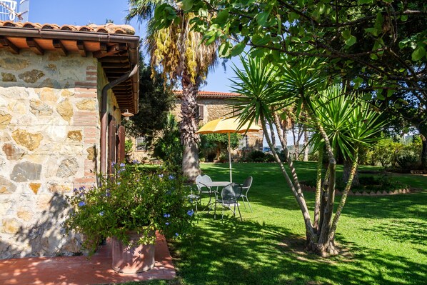 A stone building with a tiled roof, a patio with a table and chairs, and a well-maintained lawn with a palm tree.