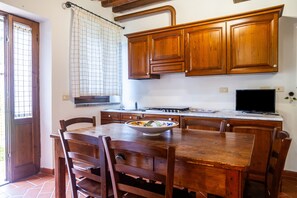 A kitchen with wooden cabinets, a dining table with chairs, a bowl on the table, and a window with a curtain.