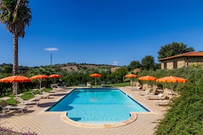 Une piscine entourée de verdure, avec des chaises longues et des parasols, sous un ciel dégagé.