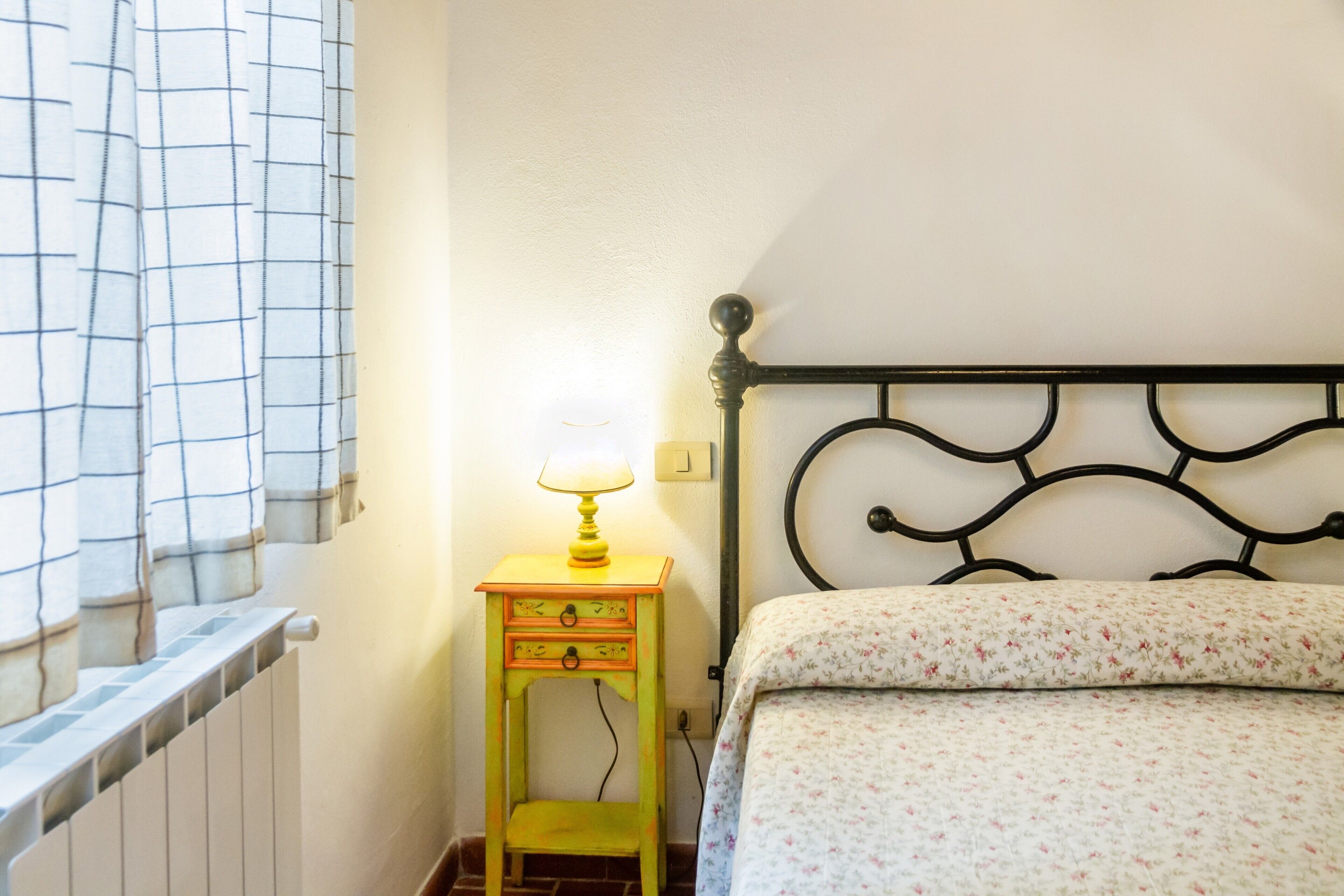 Une chambre à coucher avec un lit, une table de chevet avec une lampe, une fenêtre avec des rideaux et un radiateur.