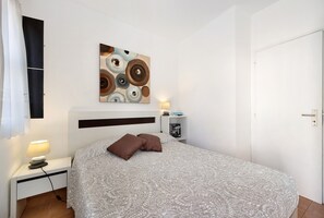 A bedroom with a bed, a bedside table with a lamp, a wall-mounted artwork, and a nightstand with a book.
