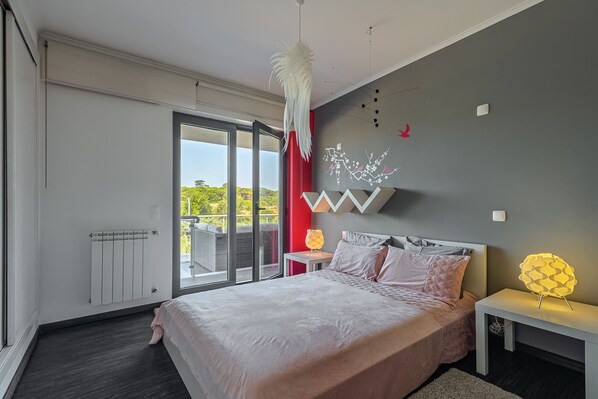 A modern bedroom with a large bed, a ceiling fan, a radiator, and a view of greenery outside. - Holiday Home 'Casa Praia Do Meco' with Private Pool, Private Terrace and Wi-Fi (Sesimbra)