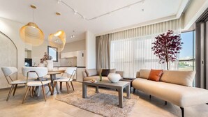 Un salon moderne avec un canapé, une table basse et des fauteuils. On y trouve également un coin repas avec une table et des chaises. Une grande fenêtre avec des rideaux est visible.