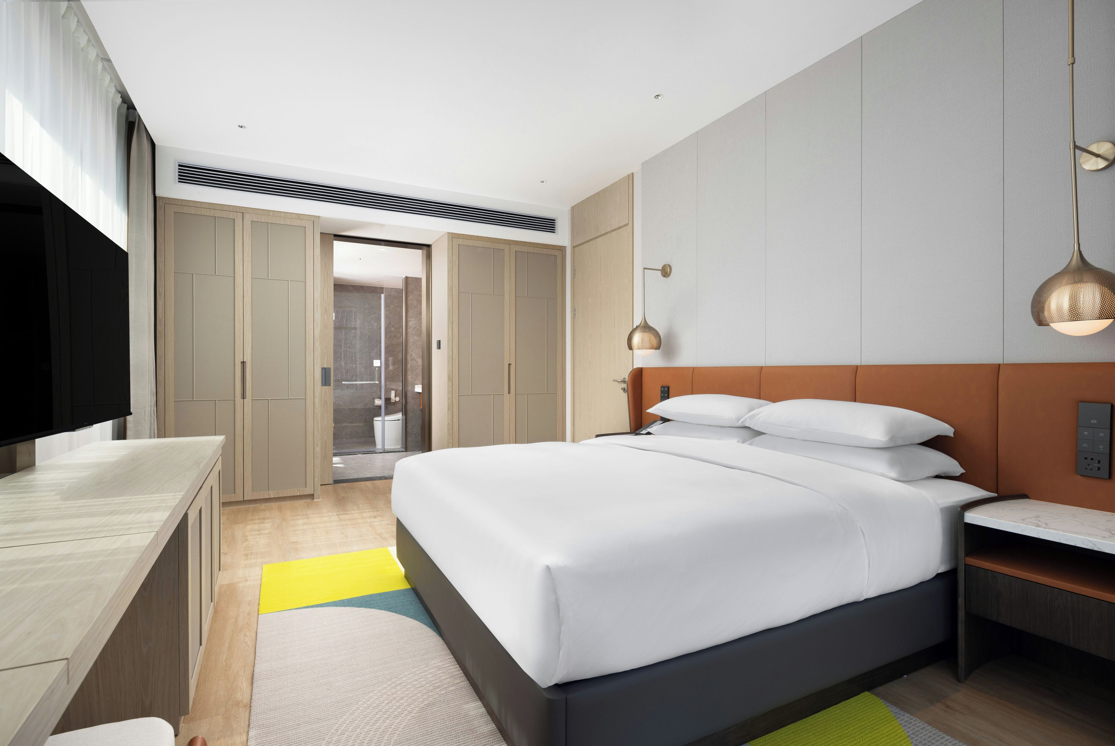 Een moderne hotelkamer met een bed, nachtkastje, televisie, garderobe en een zichtbare badkamer door een geopende deur.