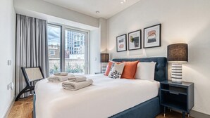 A modern hotel room with a large bed, a view of buildings through the window, and a nightstand with a lamp. - Westminster Fire Station Luxurious penthouse (London)