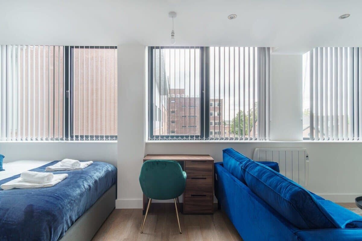 Habitació d'hotel moderna amb llit, escriptori, sofà blau i grans finestres amb persianes.