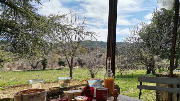 A picnic setup with bread, drinks, and a bottle on a table with a view of trees and hills.