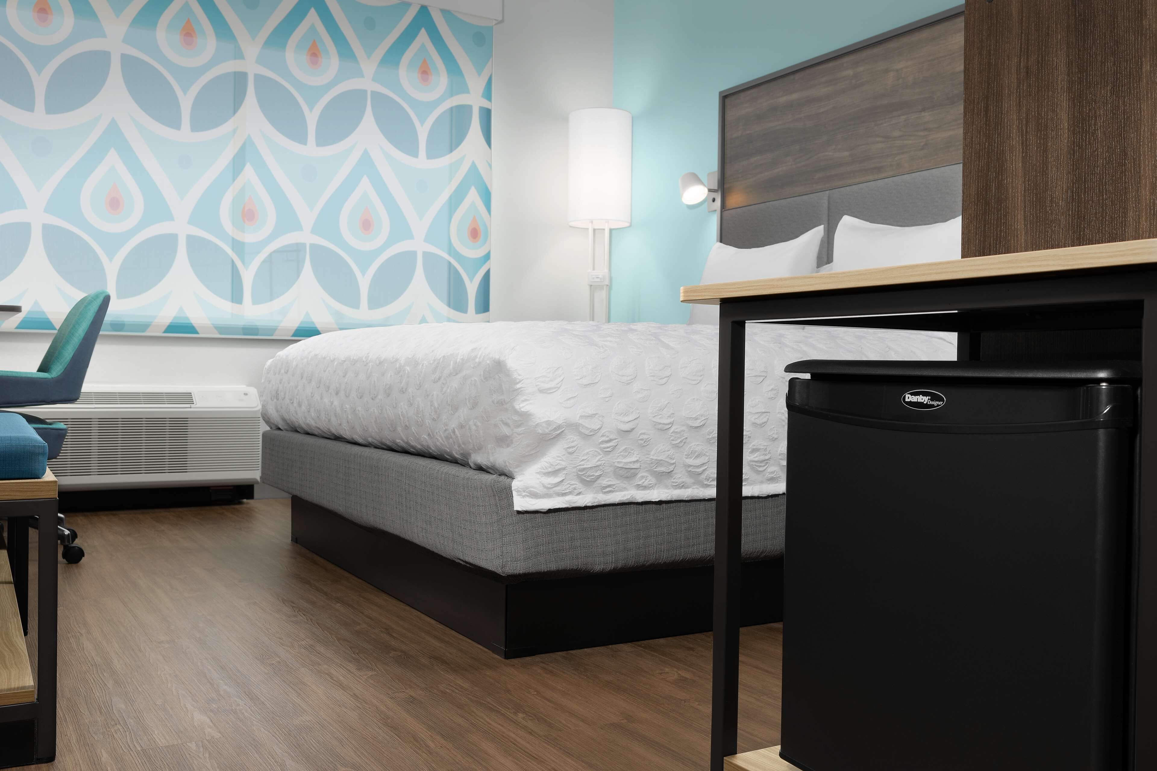 A hotel room with a bed, a desk, a chair, a lamp, and a refrigerator.