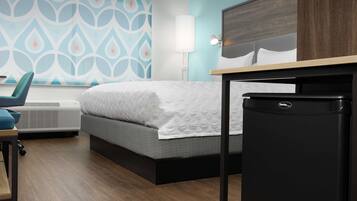 A hotel room with a bed, a desk, a chair, a lamp, and a refrigerator.