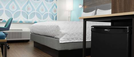 A hotel room with a bed, a desk, a chair, a lamp, and a refrigerator.