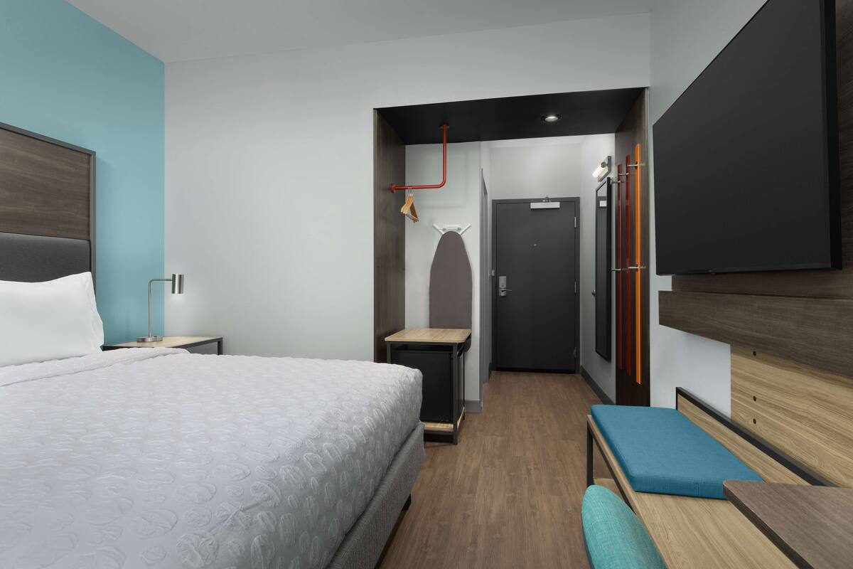 A hotel room with a large bed, a bedside table, a bench, a wardrobe, and a door leading to another room.