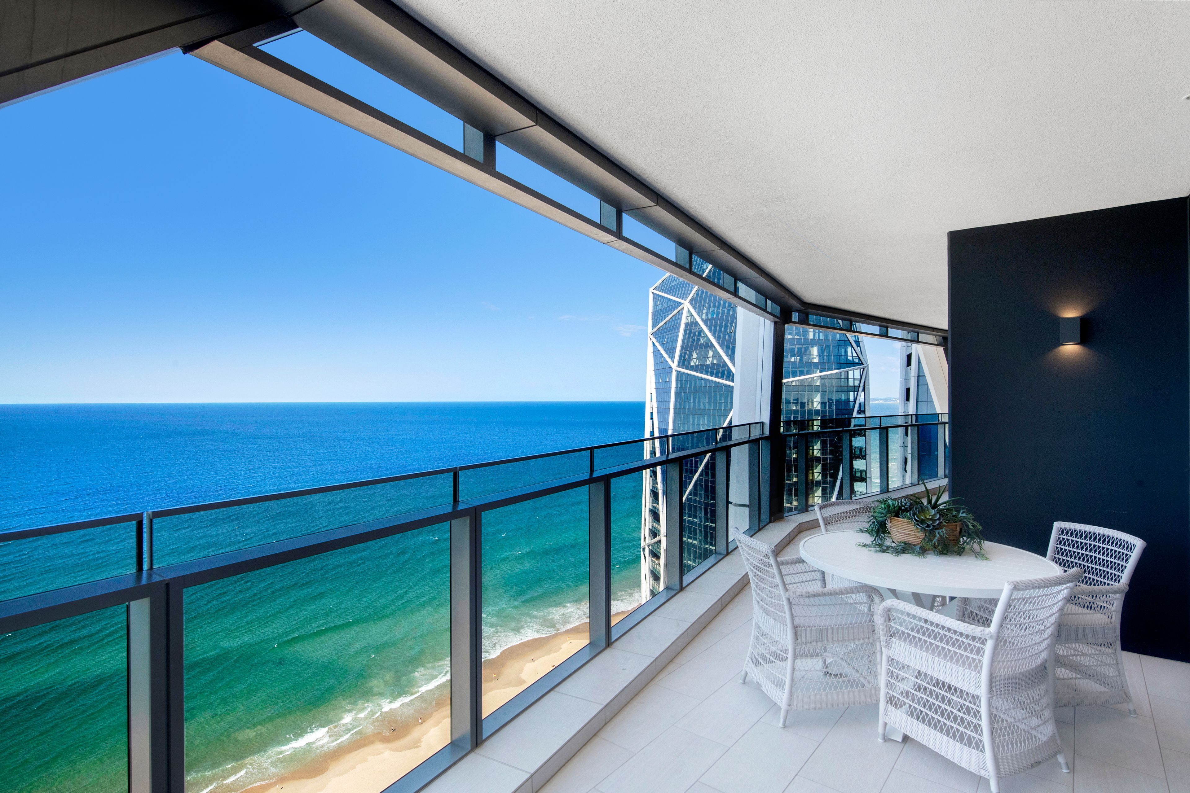 A balcony with a view of the ocean and a modern building. There is a table with chairs and a small plant.