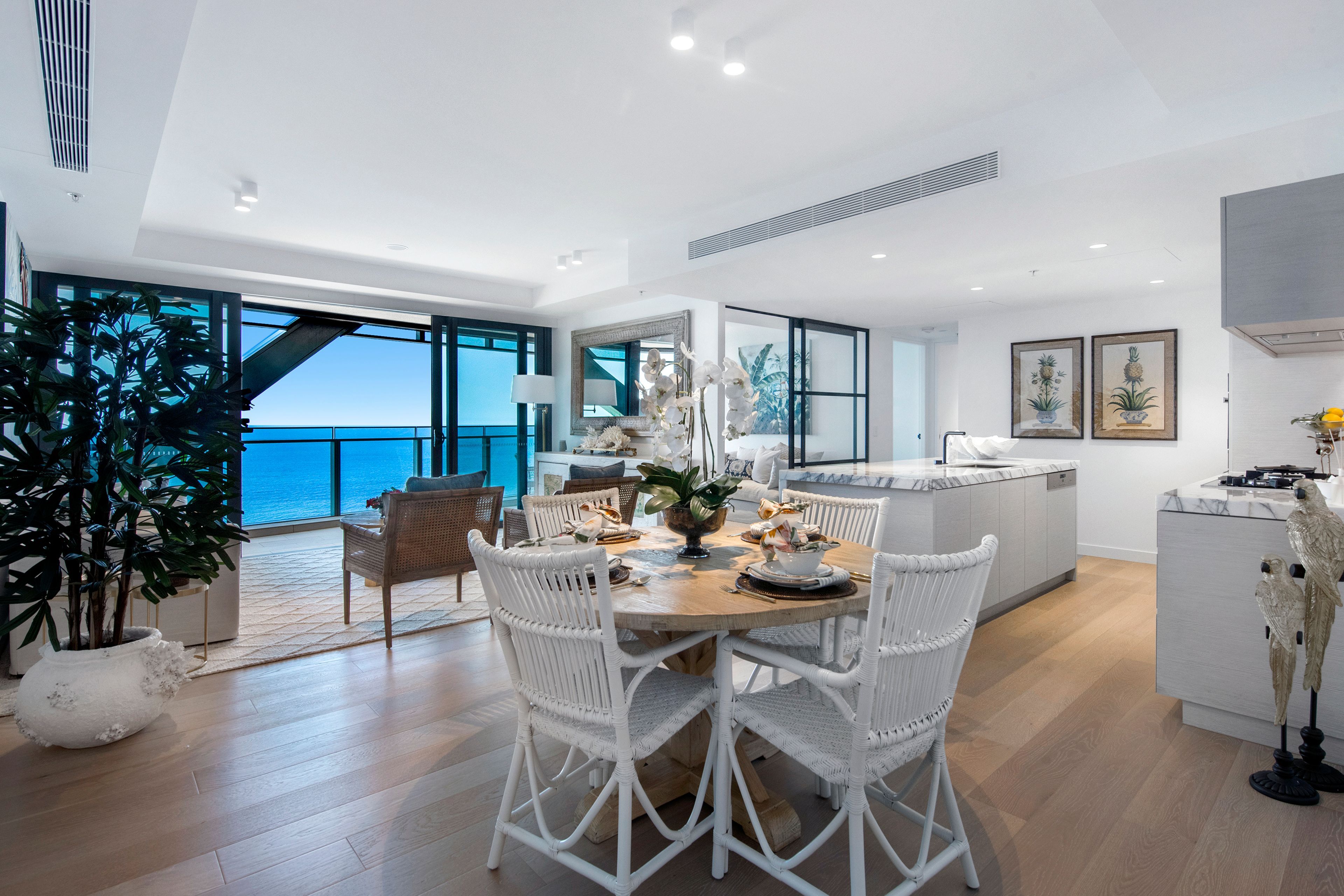 A modern kitchen with a large island, white wicker chairs, and a view of the sea.