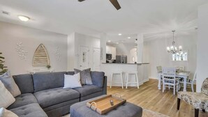 Una sala moderna con un sofá gris, una mesa de centro de madera y una zona de comedor con sillas blancas y una araña.