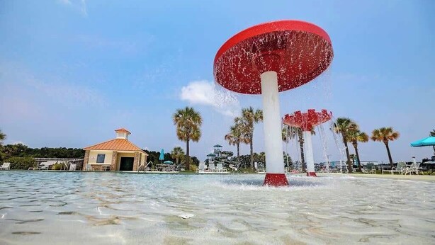 Una fuente de agua con una parte superior grande y roja, rodeada de palmeras y un pequeño edificio con techo rojo.
