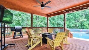 A covered deck with wooden flooring, yellow Adirondack chairs, a grill, a TV, and a pool in the background. - Beautifully decorated and spacious home with a pool. (Rex)