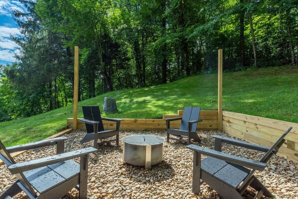 Un espacio exterior con cuatro sillas Adirondack, una mesa redonda y un brasero, todo rodeado por una terraza de madera y grava.