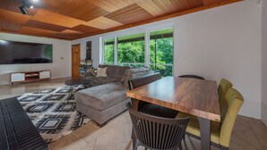 Un soggiorno moderno con soffitto in legno, una grande televisione, un divano, un tavolo da pranzo e vista sul verde.