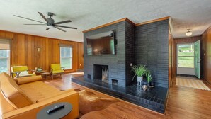 Un soggiorno moderno con camino, pavimento in legno, ventilatore a soffitto e una grande finestra.