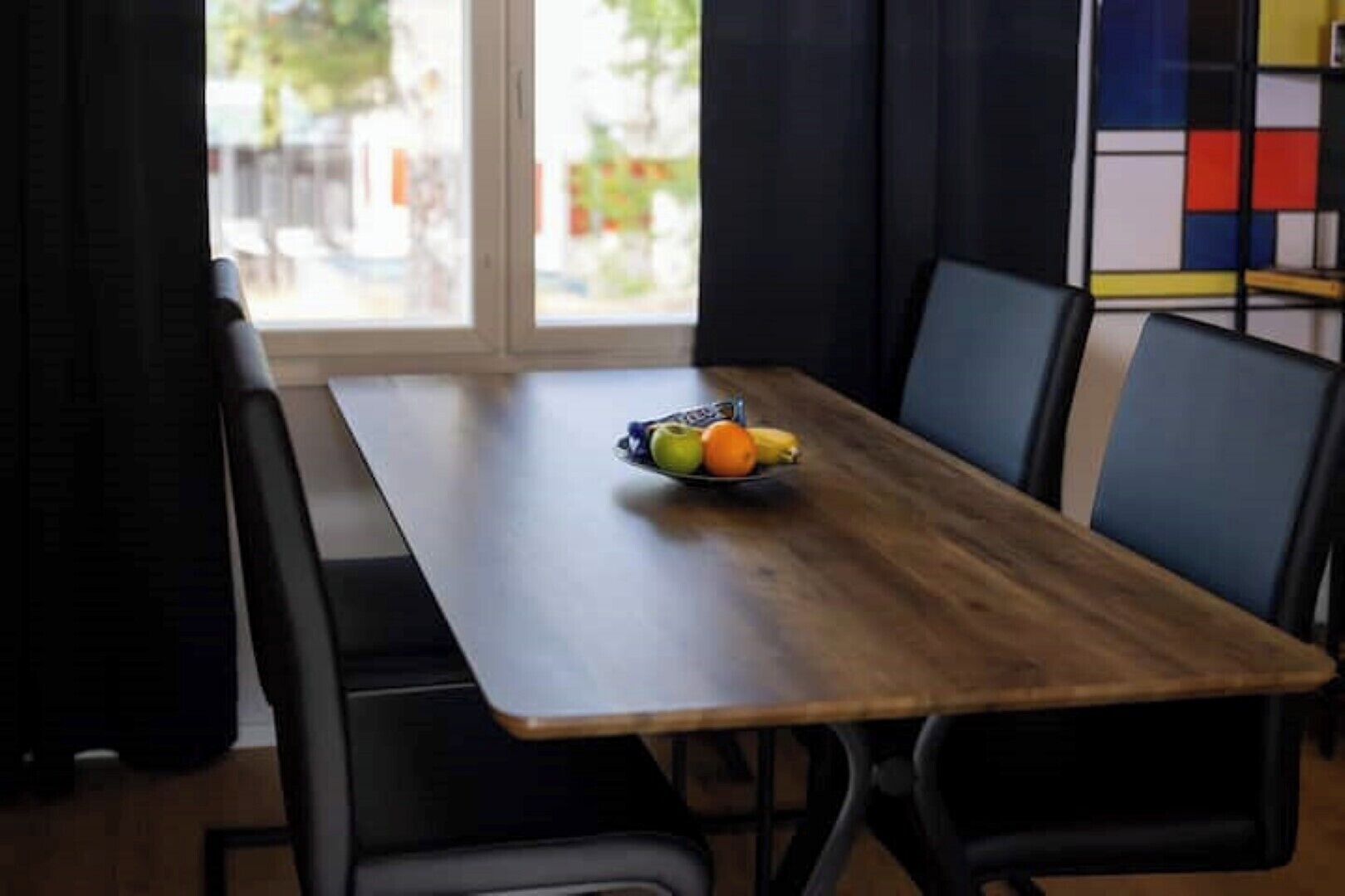 A dining table with a fruit bowl, black chairs, and a view of the outdoors through a window.