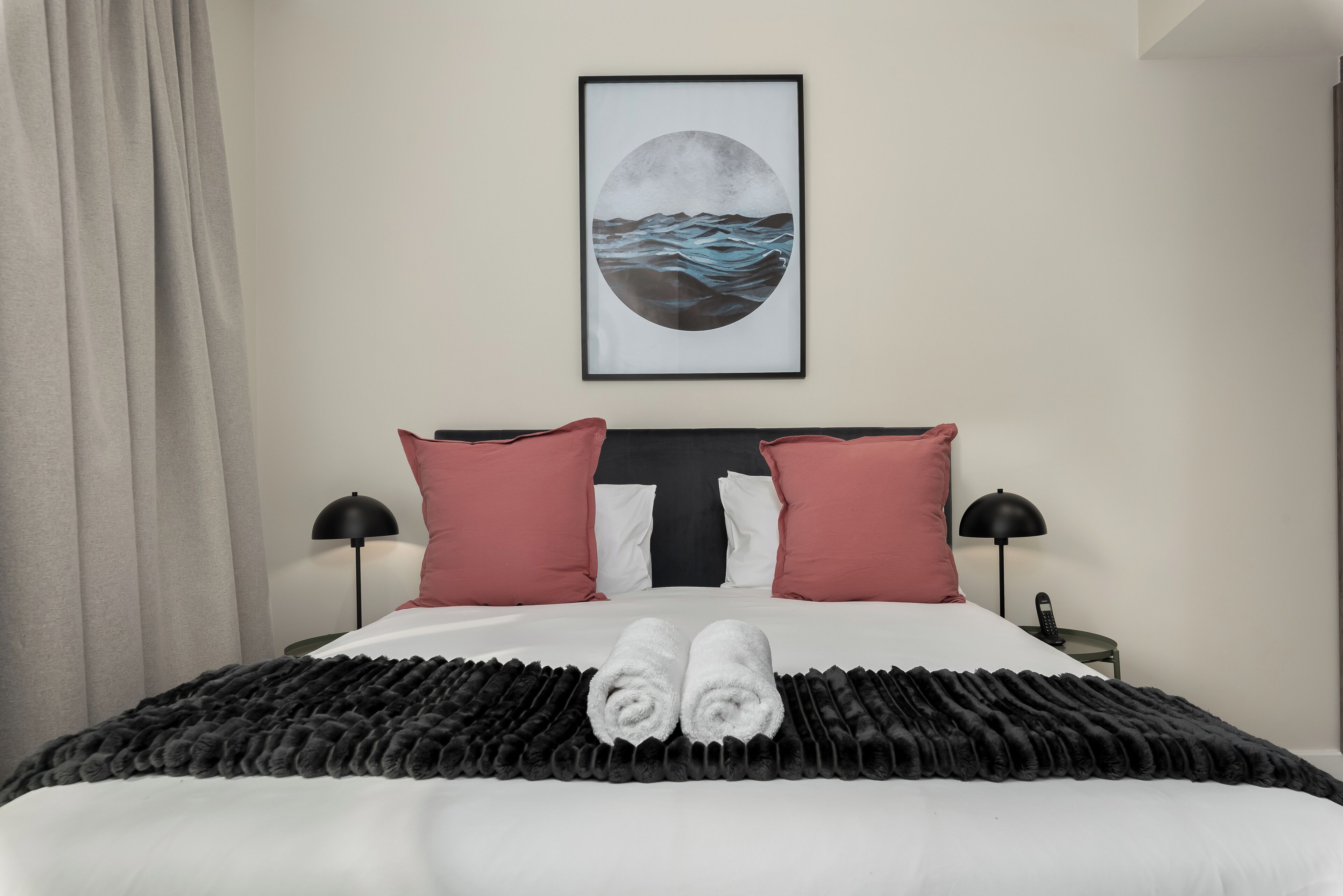 Una cama bien hecha con edredón blanco y negro, dos almohadas rojas y un cuadro enmarcado encima.