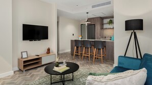 Uma sala de estar moderna com televisão de tela plana, uma mesa de centro com livros e um vaso, um sofá azul e uma área de cozinha com banquetas.