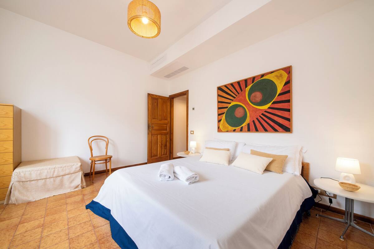 A hotel room with a large bed, a wooden chair, a small table with a lamp, and a painting on the wall.
