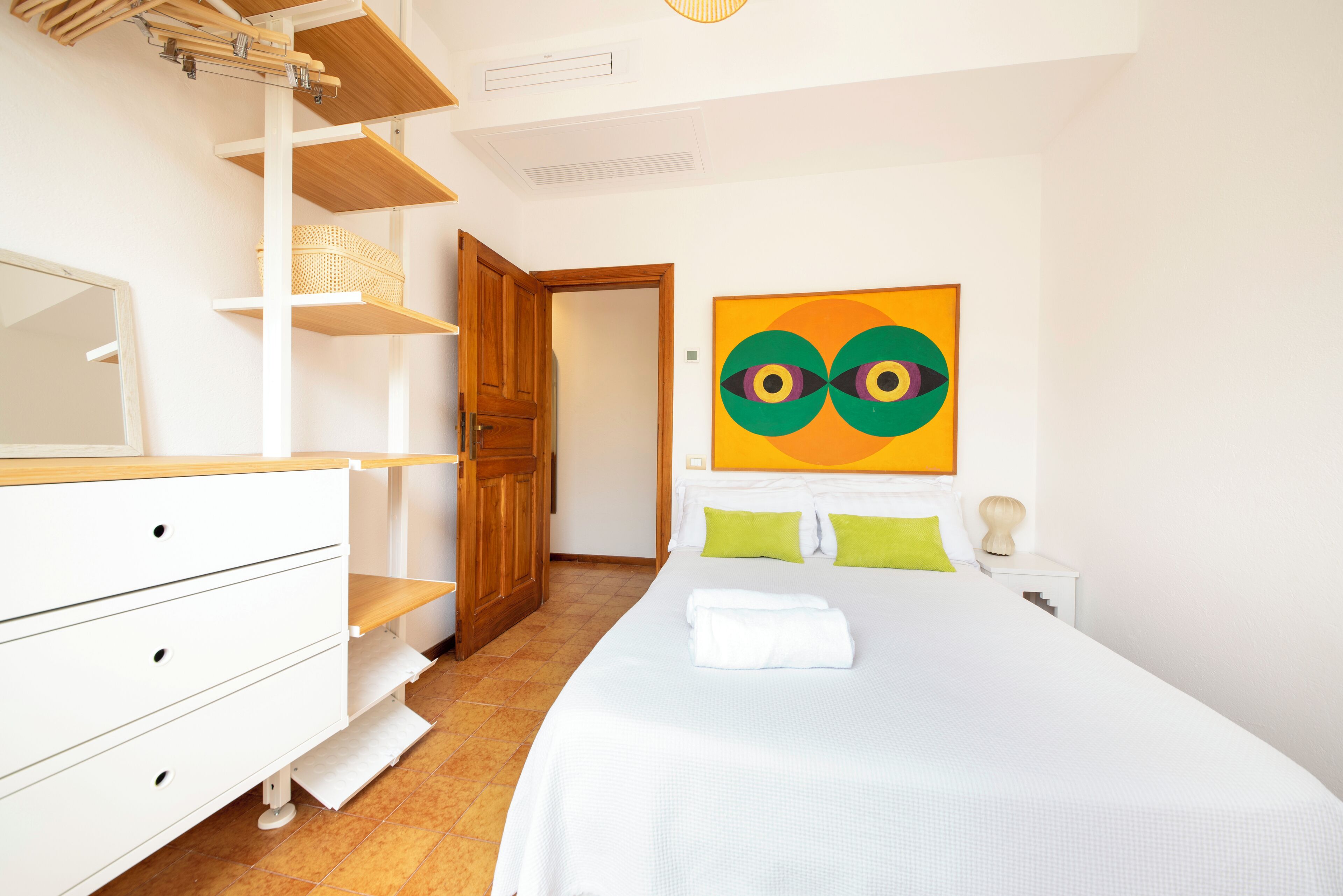 A bedroom with a bed, a dresser, a wooden door, a wall-mounted shelf, and a colorful abstract painting.