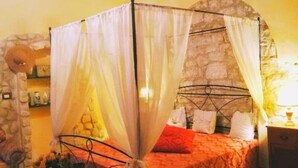 Una cama con dosel, un muro de piedra, una araña y una mesita de noche con lámpara.