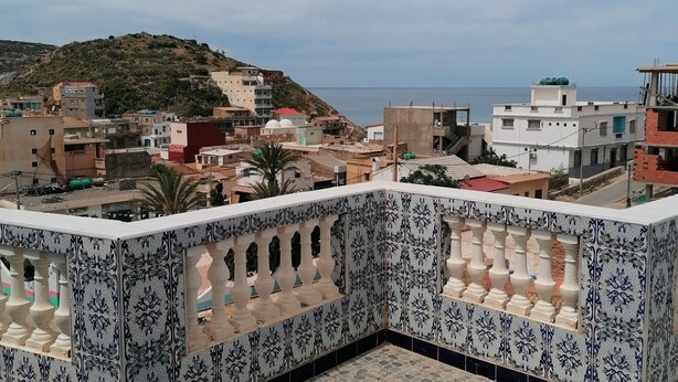 A rooftop terrace with patterned tiles, a view of the sea, and buildings in the background. - Bouzedjar-mer (Bou Zadjar)