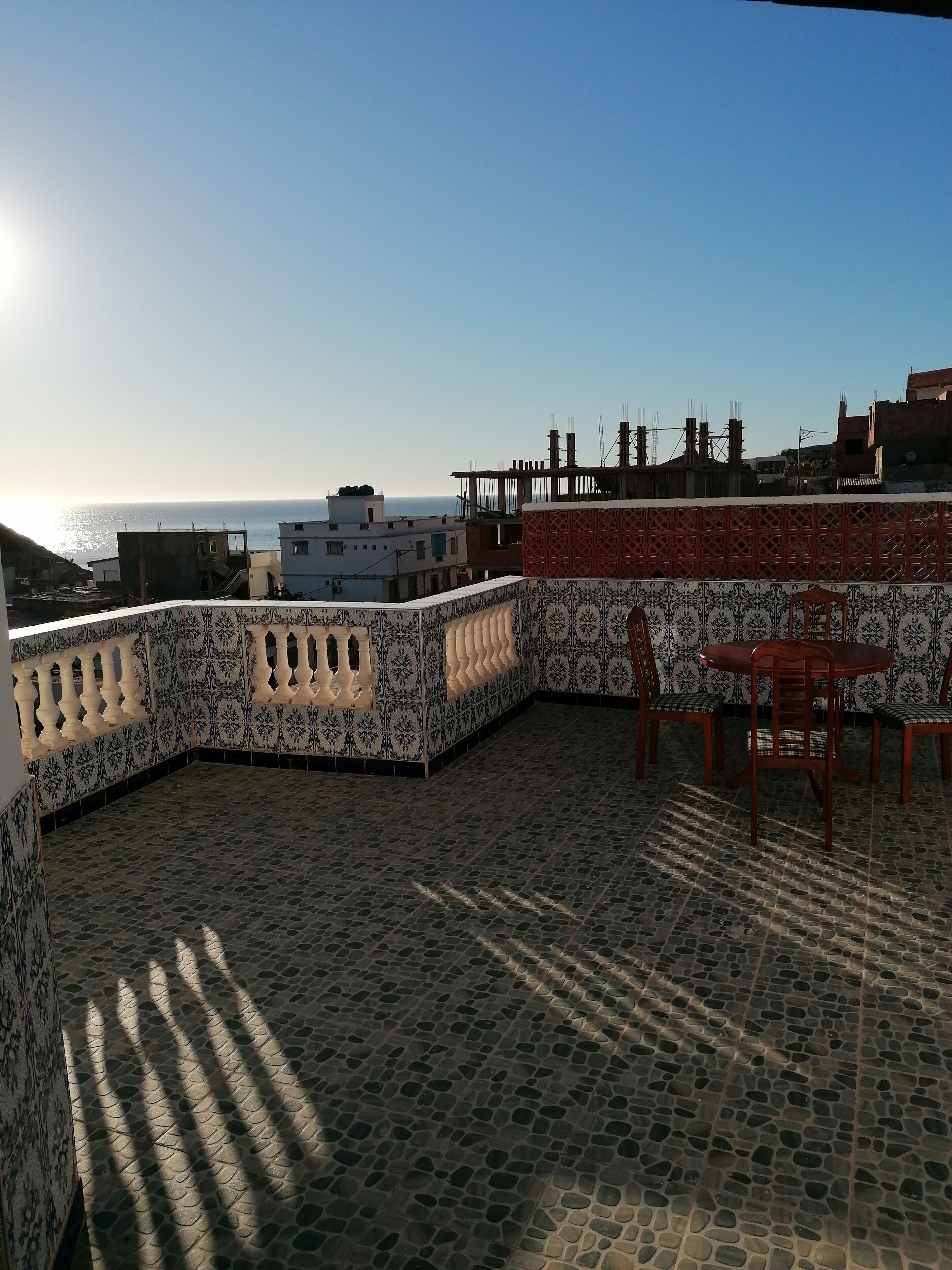 Eine Dachterrasse mit gemusterten Fliesen, einem Esstisch mit Stühlen und Blick auf das Meer und Gebäude im Hintergrund.