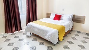 A neatly made bed with a white and yellow bedspread, a red pillow, and a wooden headboard. The room has a geometric patterned floor and large windows with brown curtains. - Mang Hotel (Nairobi)