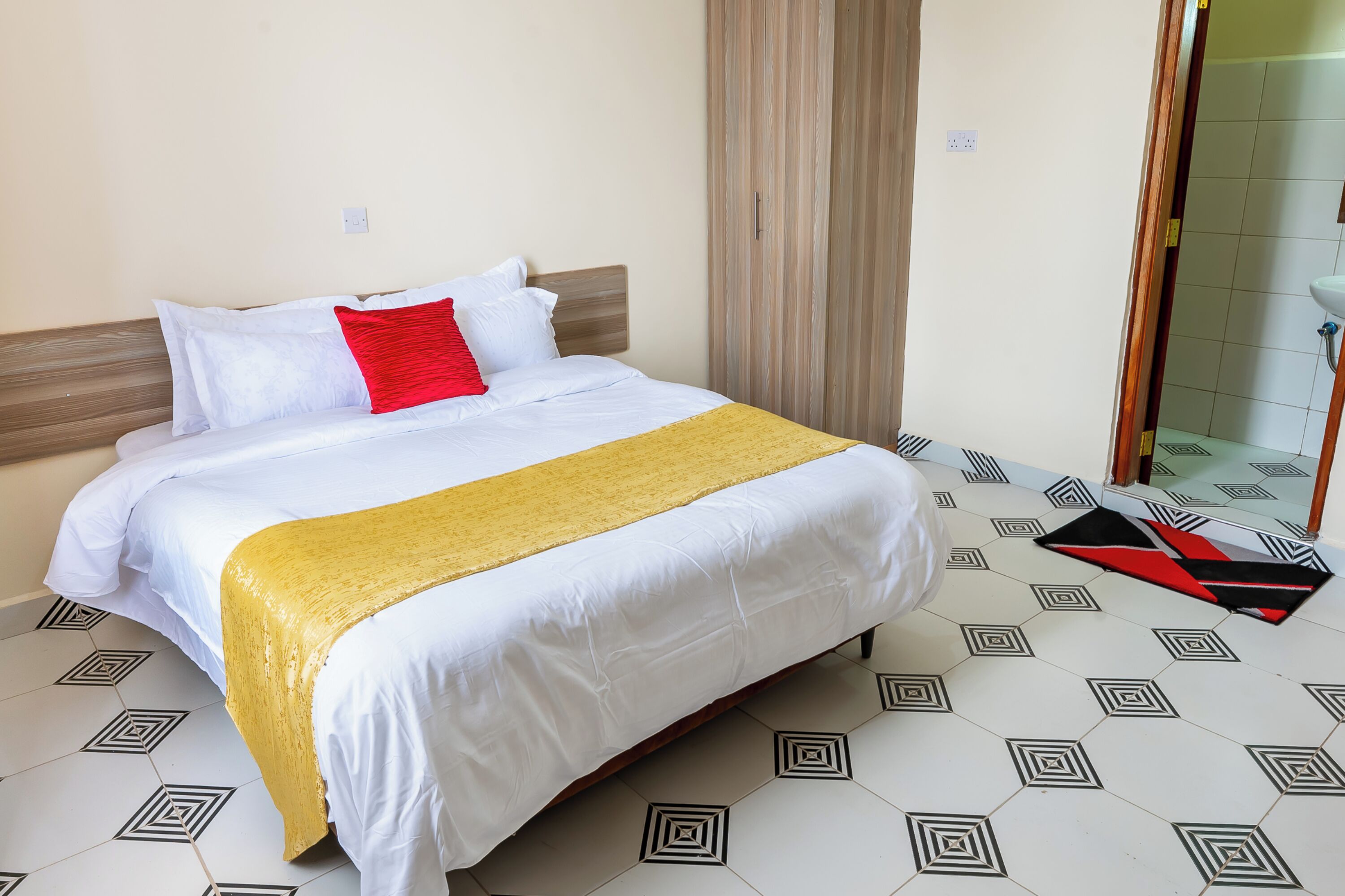 Habitación de hotel con una cama, una manta amarilla, una almohada roja y un suelo con estampado geométrico.