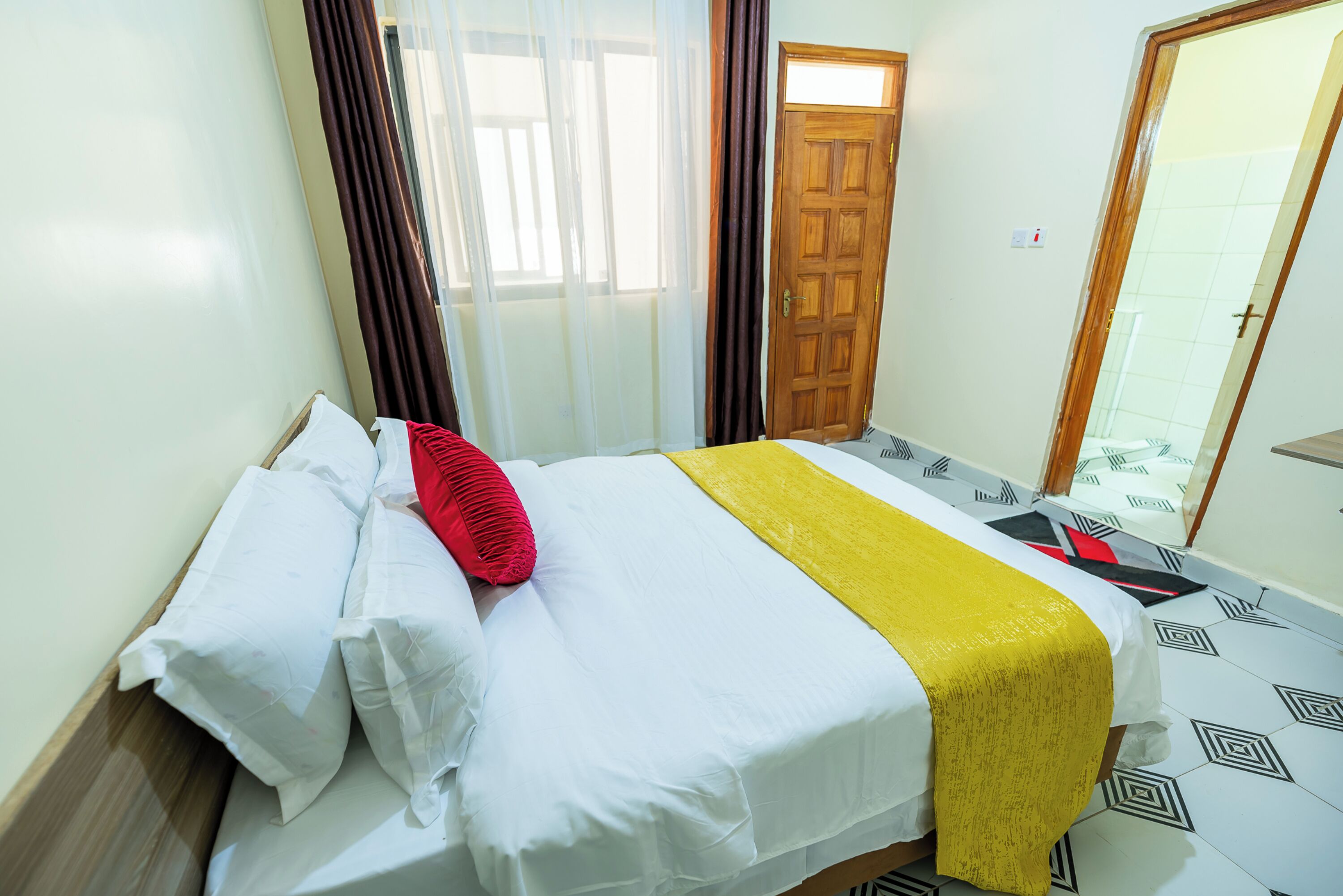 A neatly made bed with white and yellow linens, a red pillow, and a wooden headboard.