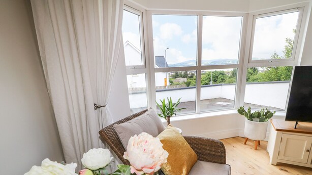 A room with a wicker sofa, a television on a wooden cabinet, and a view of a green landscape through large windows. - 12 Hillview (Buncrana)