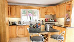 A modern kitchen with wooden cabinets, a central island with bar stools, a wine rack, and a window with a view of greenery. - 12 Hillview (Buncrana)