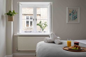 A bedroom with a bed, a window with a view of a brick building, a radiator, a plant, and a breakfast tray with toast and juice.