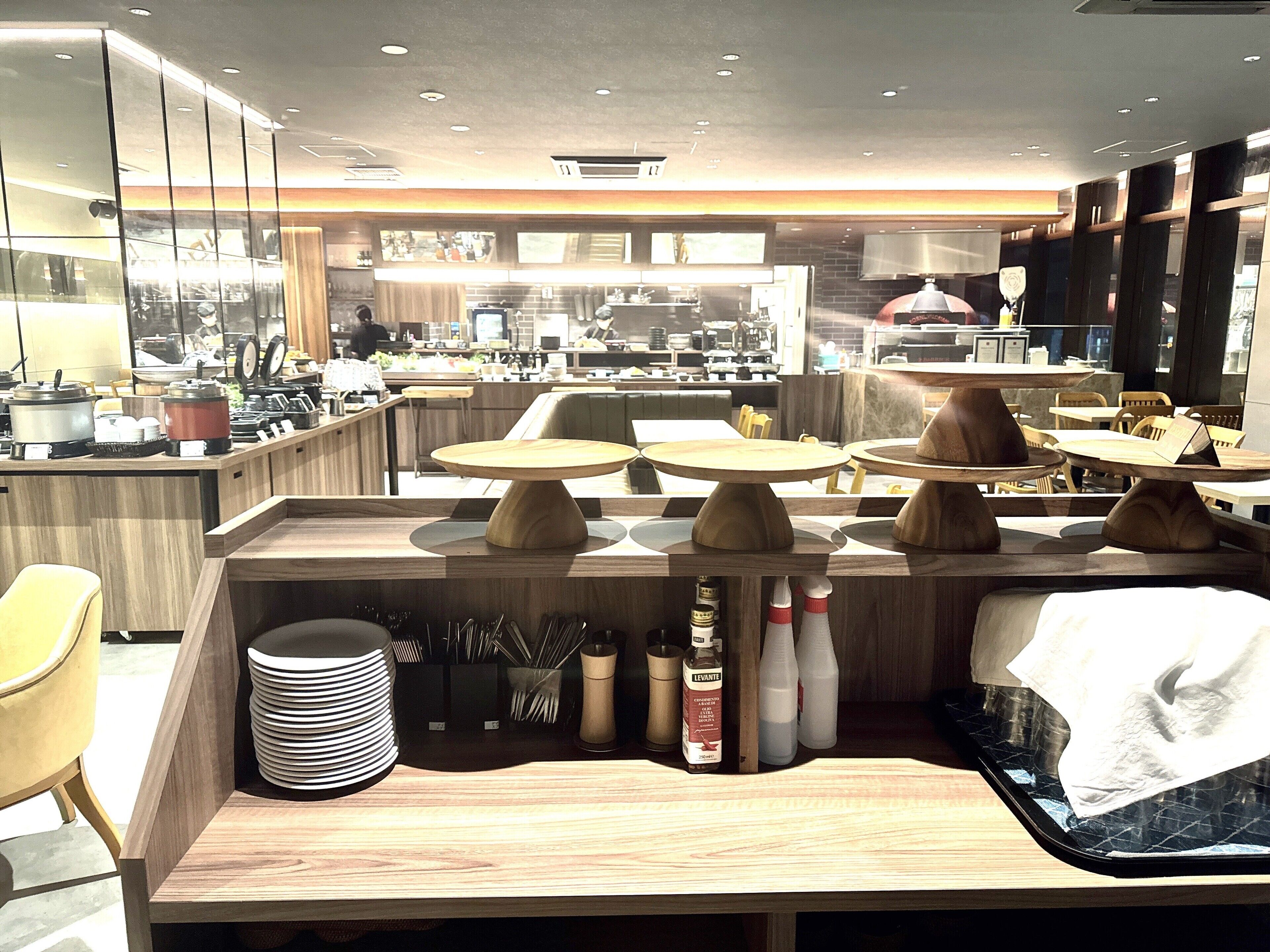 A modern restaurant interior with wooden tables, a kitchen area in the background, and various dining utensils on the counter.