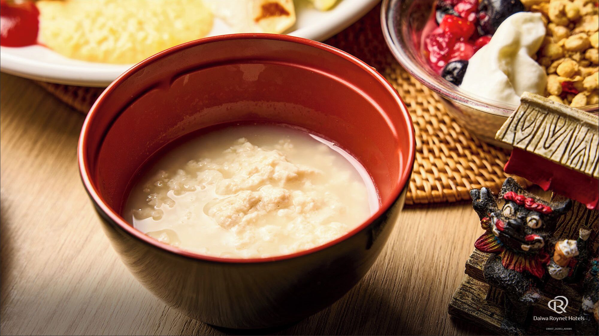 A bowl of rice porridge in a red bowl, a plate with yellow food, and a bowl of berries and nuts.