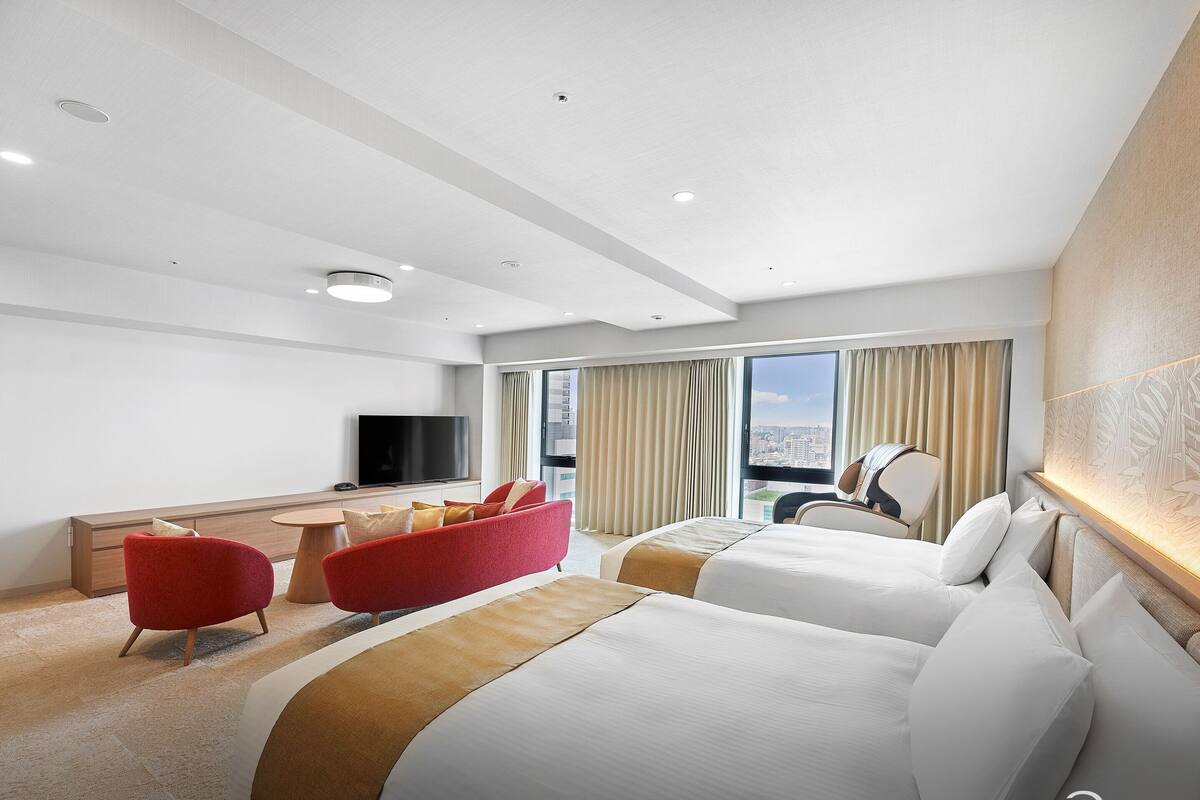 Een hotelkamer met twee bedden, een rode bank, een bureau met televisie en uitzicht over de stad.