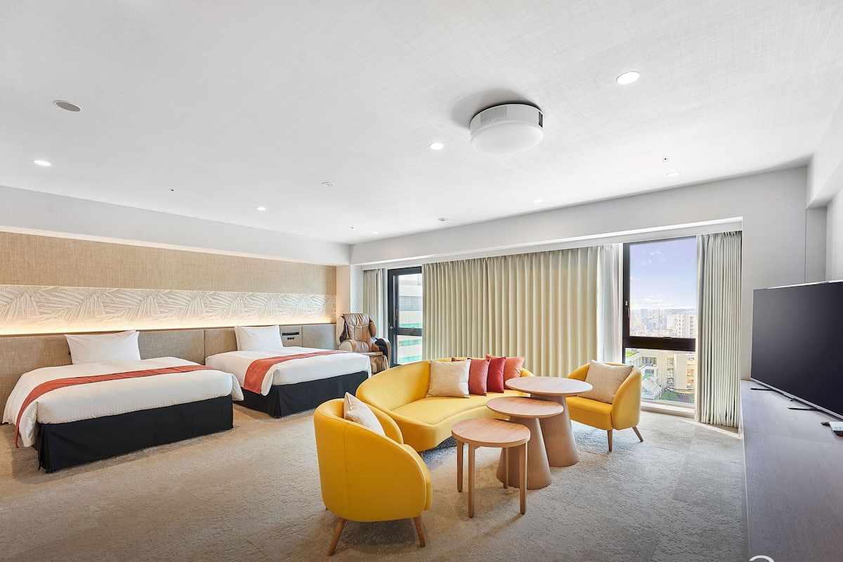 Een hotelkamer met twee bedden, een bank, een televisie en een groot raam met uitzicht op de stad.