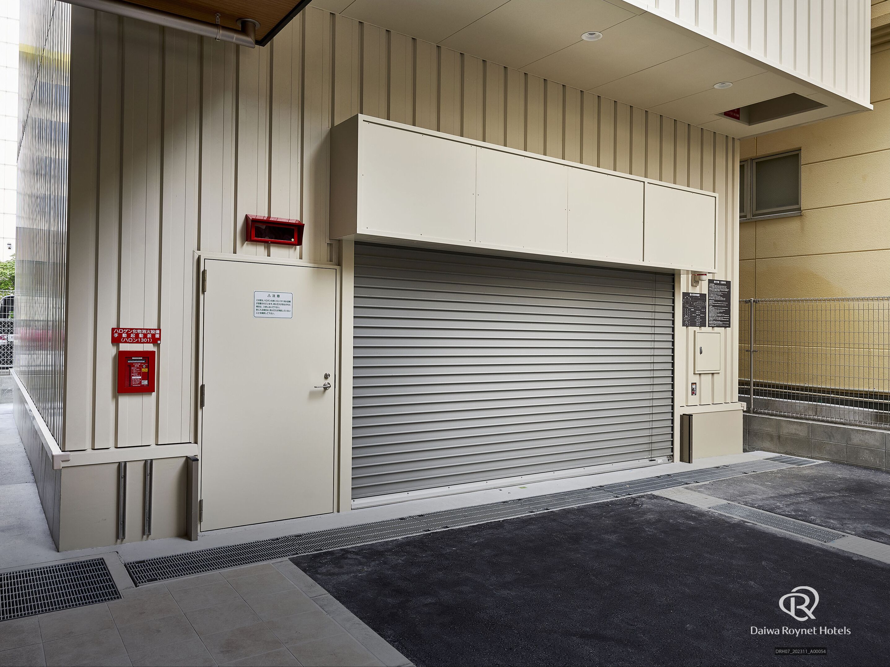A hotel entrance with a closed metal shutter door, a fire alarm box, and a security camera.