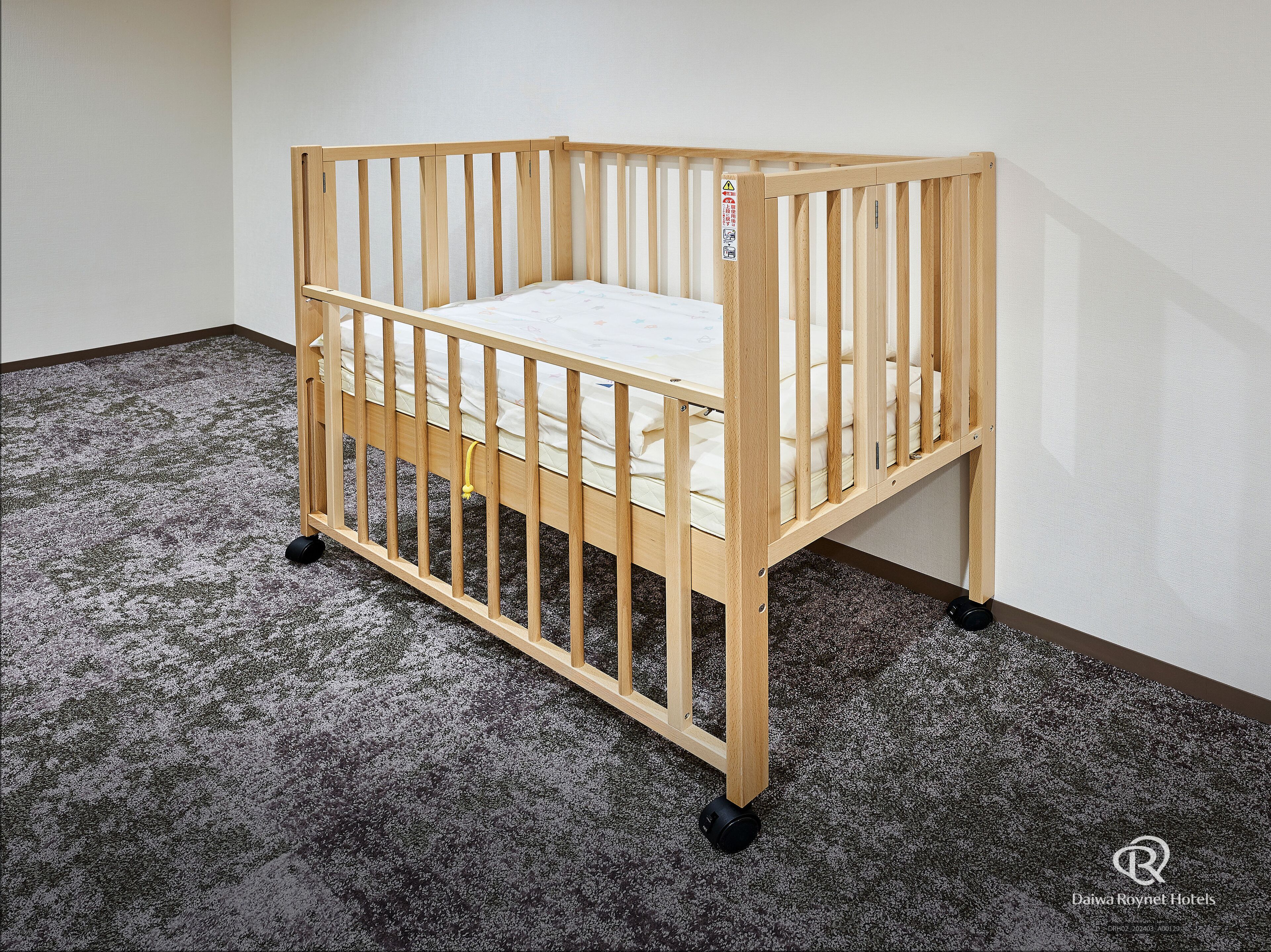 Una cuna de madera con colchón, ruedas y una etiqueta de seguridad en el lateral, sobre un piso alfombrado en una habitación con paredes lisas.