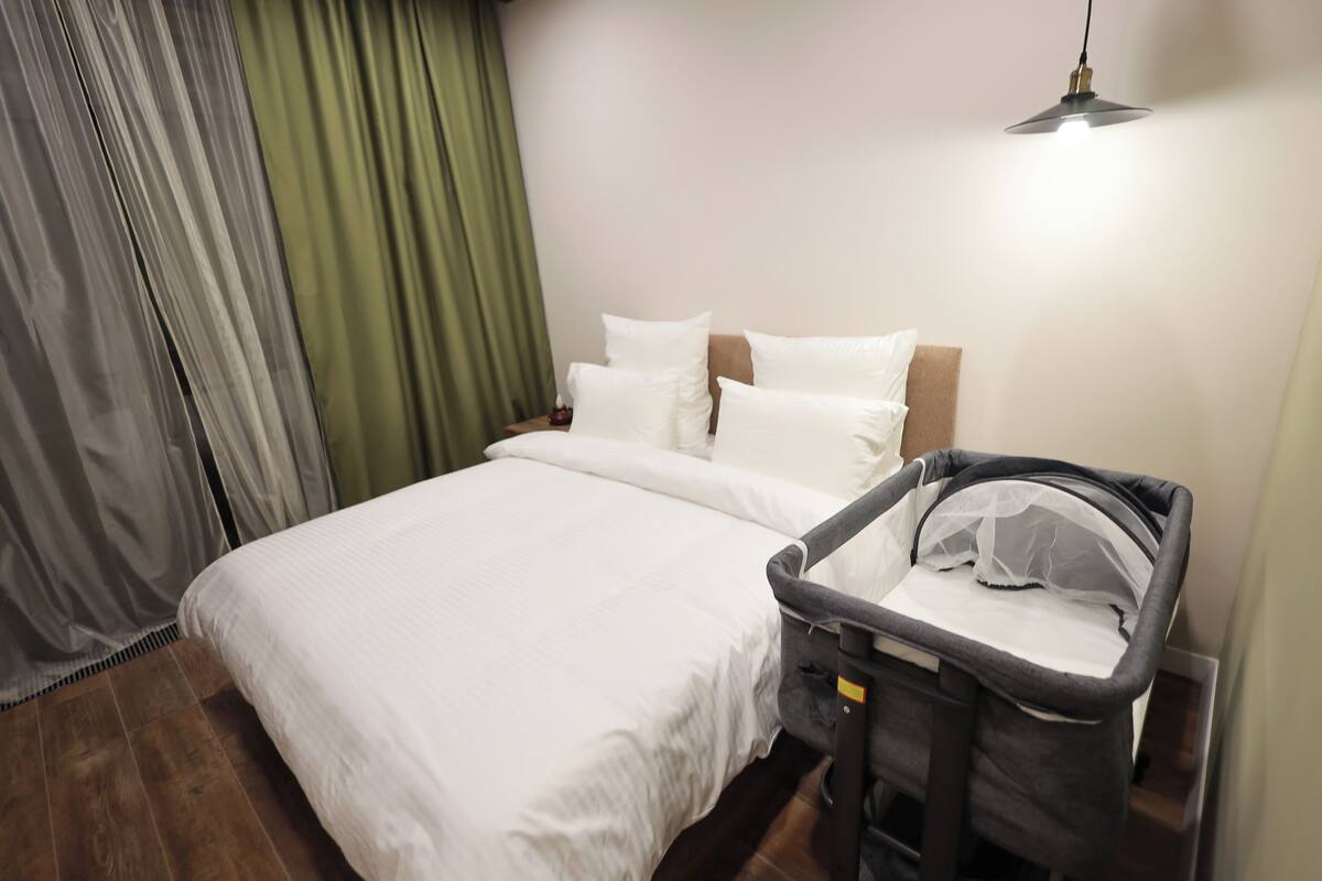 Una habitación de hotel con una cama, una cuna, una cortina verde y una lámpara colgante.