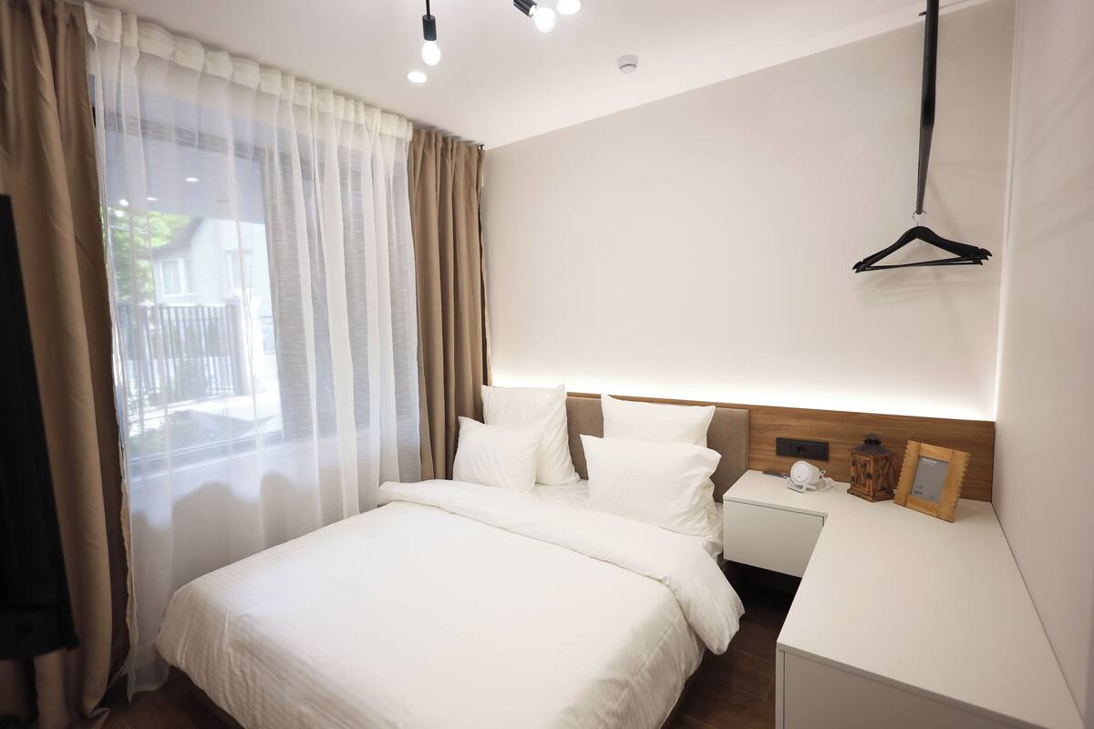 Habitación de hotel con una cama grande, una mesita de noche con lámpara y vistas al exterior a través de cortinas traslúcidas.